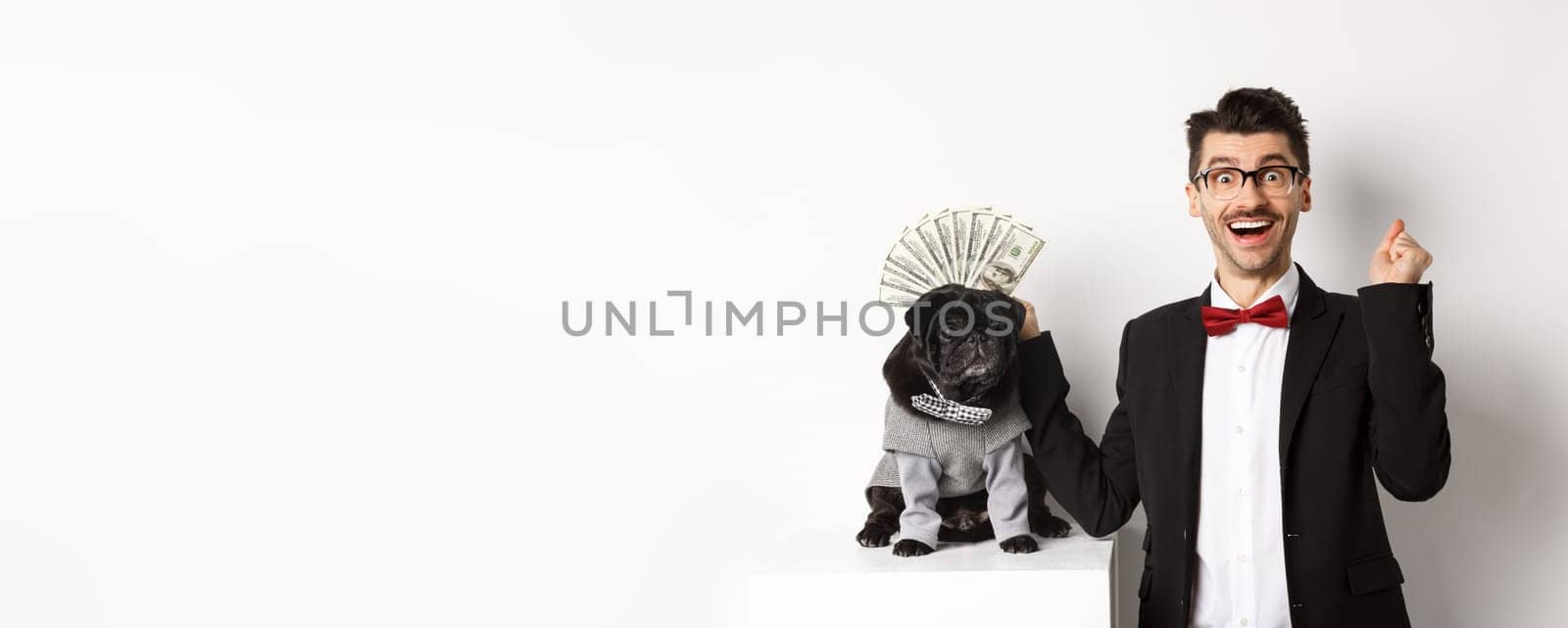 Happy man winning money, wearing costume and showing dollars near his cute black dog in suit, standing over white background.