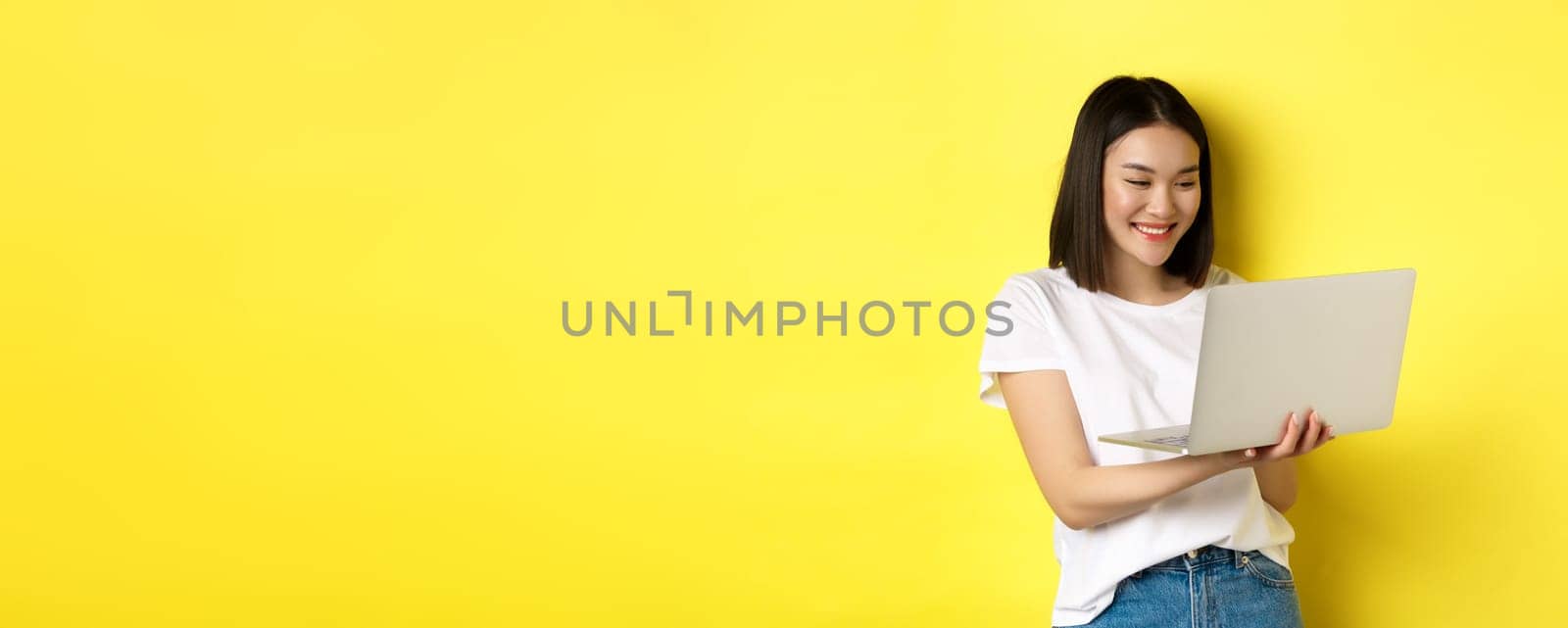 Cute asian female student working on laptop, reading screen and smiling, yellow background.