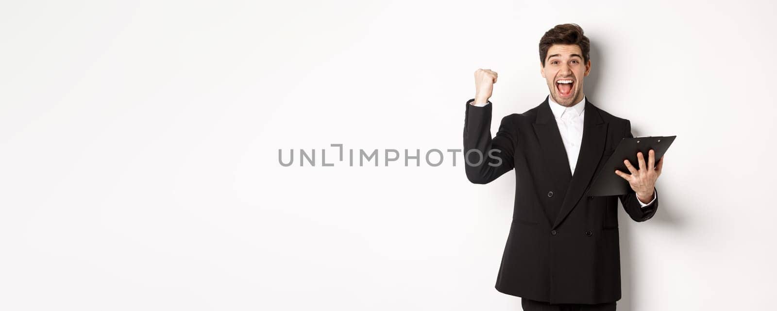 Portrait of excited handsome businessman in black suit, holding clipboard and making fist pump, achieve goal and rejoicing, standing over white background.