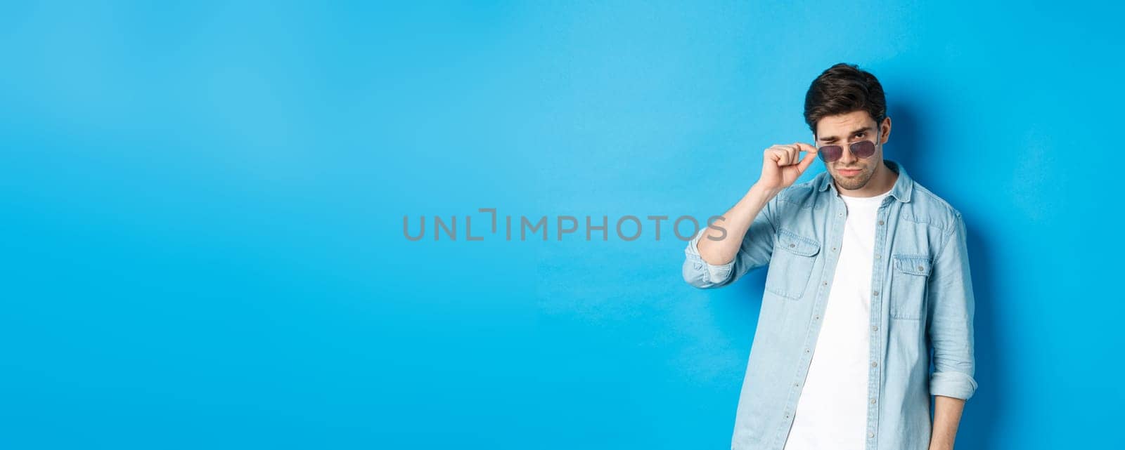 Confident macho man put on sunglasses, looking cool and sassy, standing over blue background by Benzoix