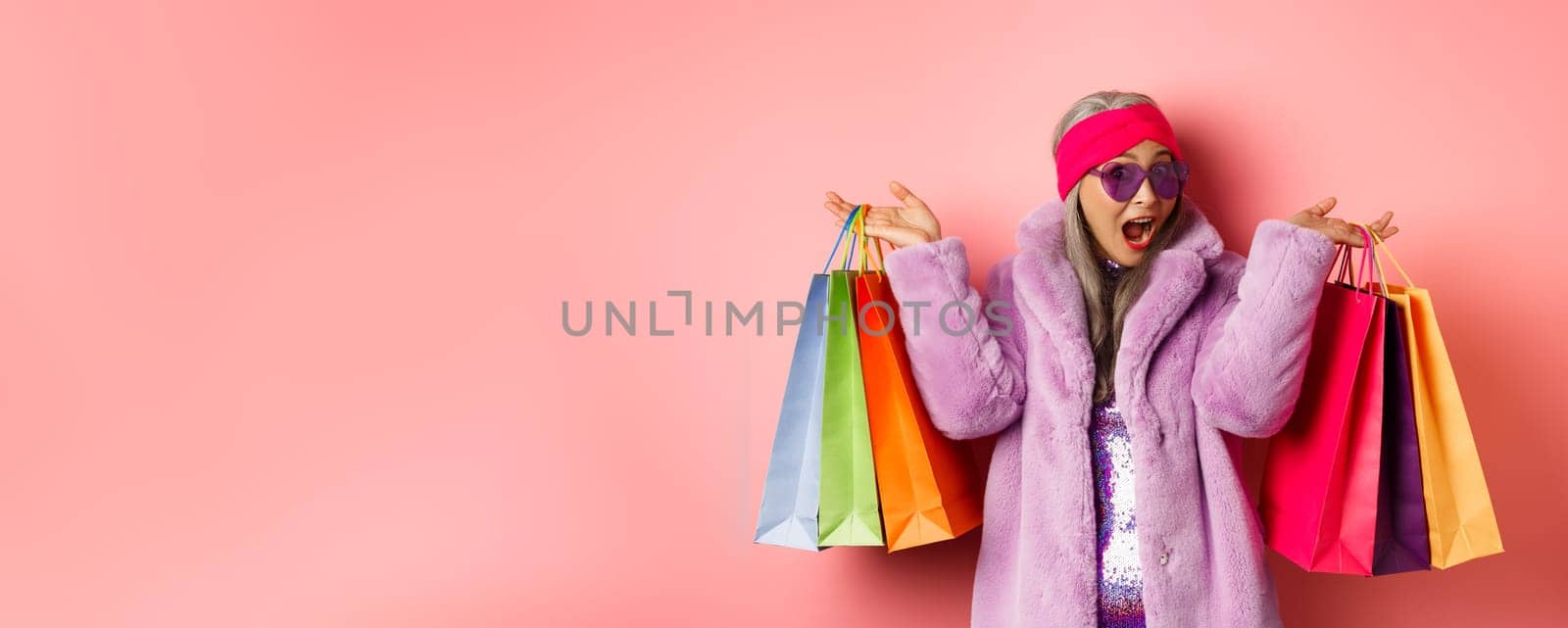 Stylish asian senior woman going shopping, wearing trendy clothes and sunglasses, holding store bags with gifts, treat yourself concept, pink background.