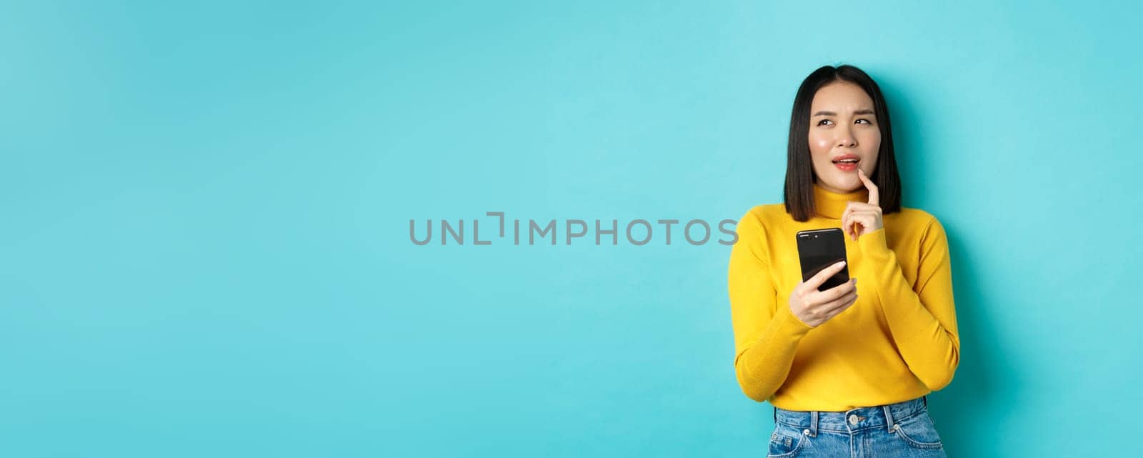 Pensive asian girl holding smartphone and thinking what to order online, standing over blue background.