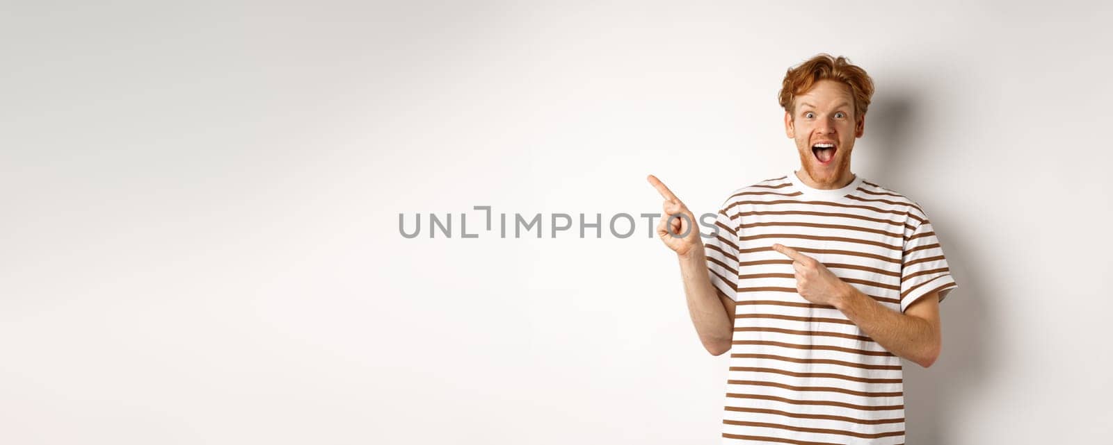 Cheerful man with red hair checking out promo offfer, pointing left at logo and smiling at camera, standing over white background.