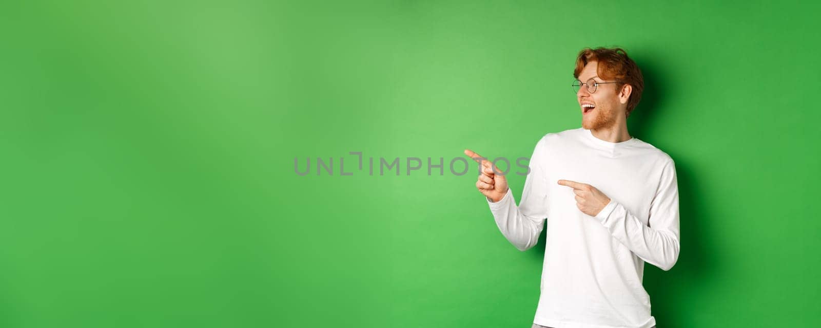 Surprised redhead man in glasses checking out promo, pointing and looking left with amazed face, standing in long-sleeve t-shirt over green background by Benzoix