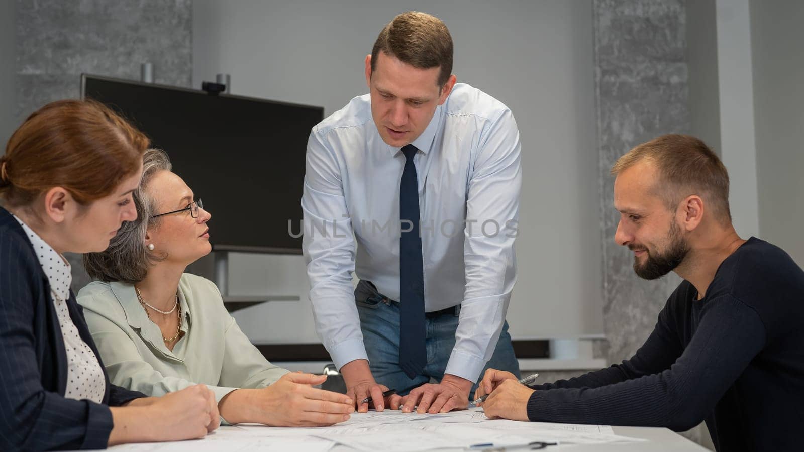 A caucasian man is standing making changes to a drawing, three colleagues are sitting at a table and listening to him