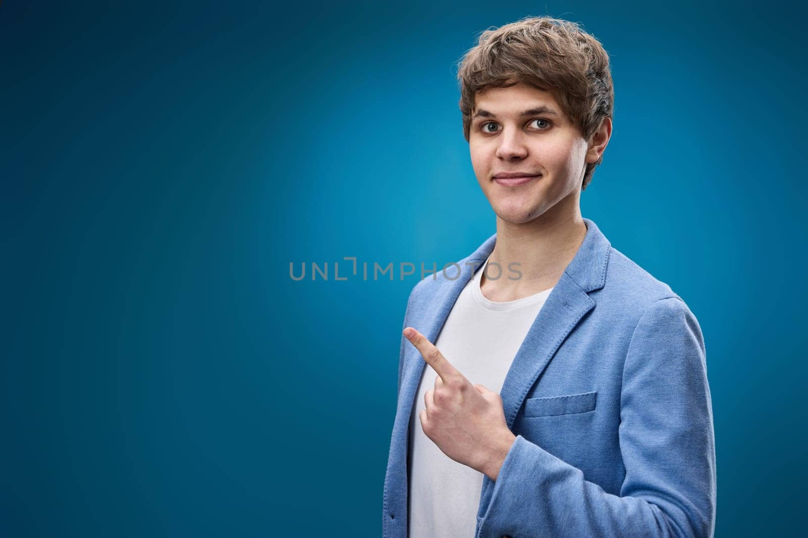 handsome caucasian young man in jacket pointing finger at empty space for advertisement on blue background