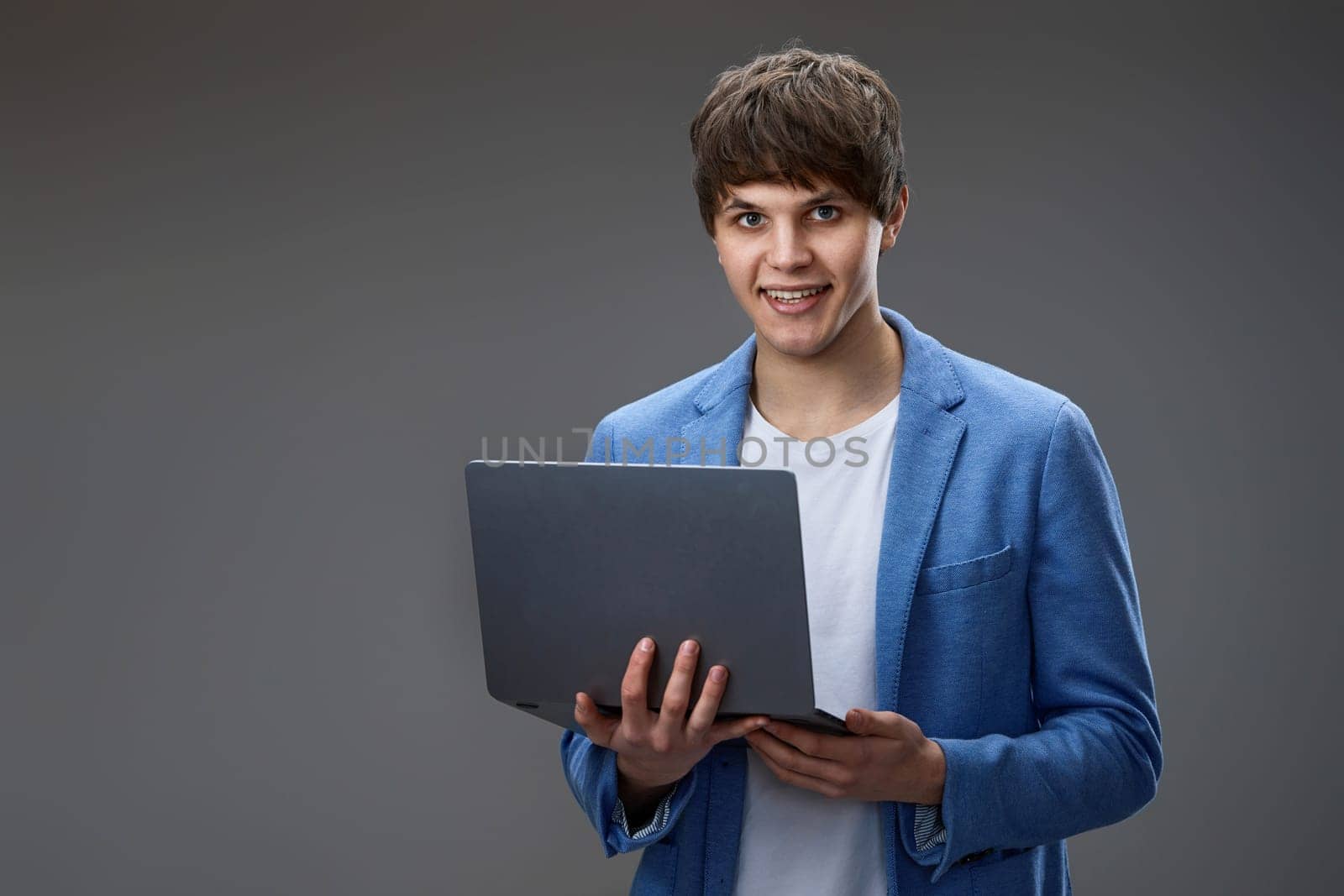 smiling caucasian young businessman in blue jacket using laptop and looking at camera isolated on gray background