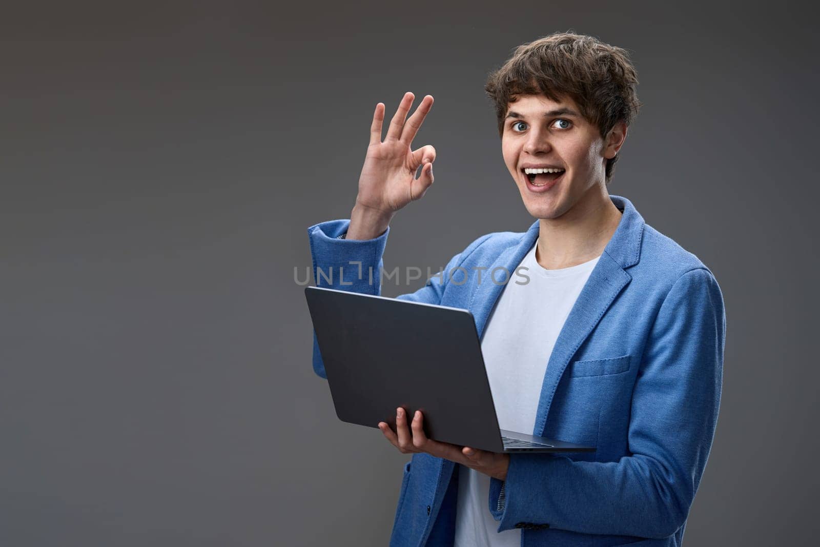 happy caucasian young guy in blue jacket using laptop and showing Ok gesture isolated on gray background. websurfing