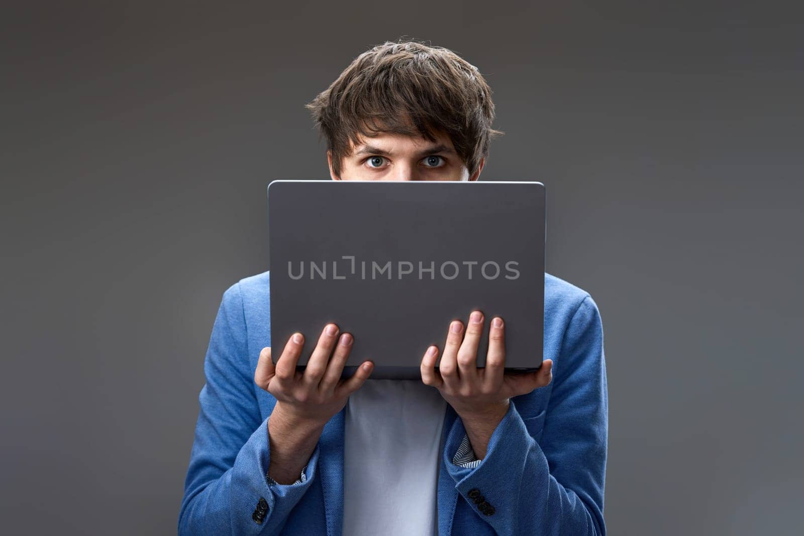 caucasian young guy hiding his face with laptop on gray background