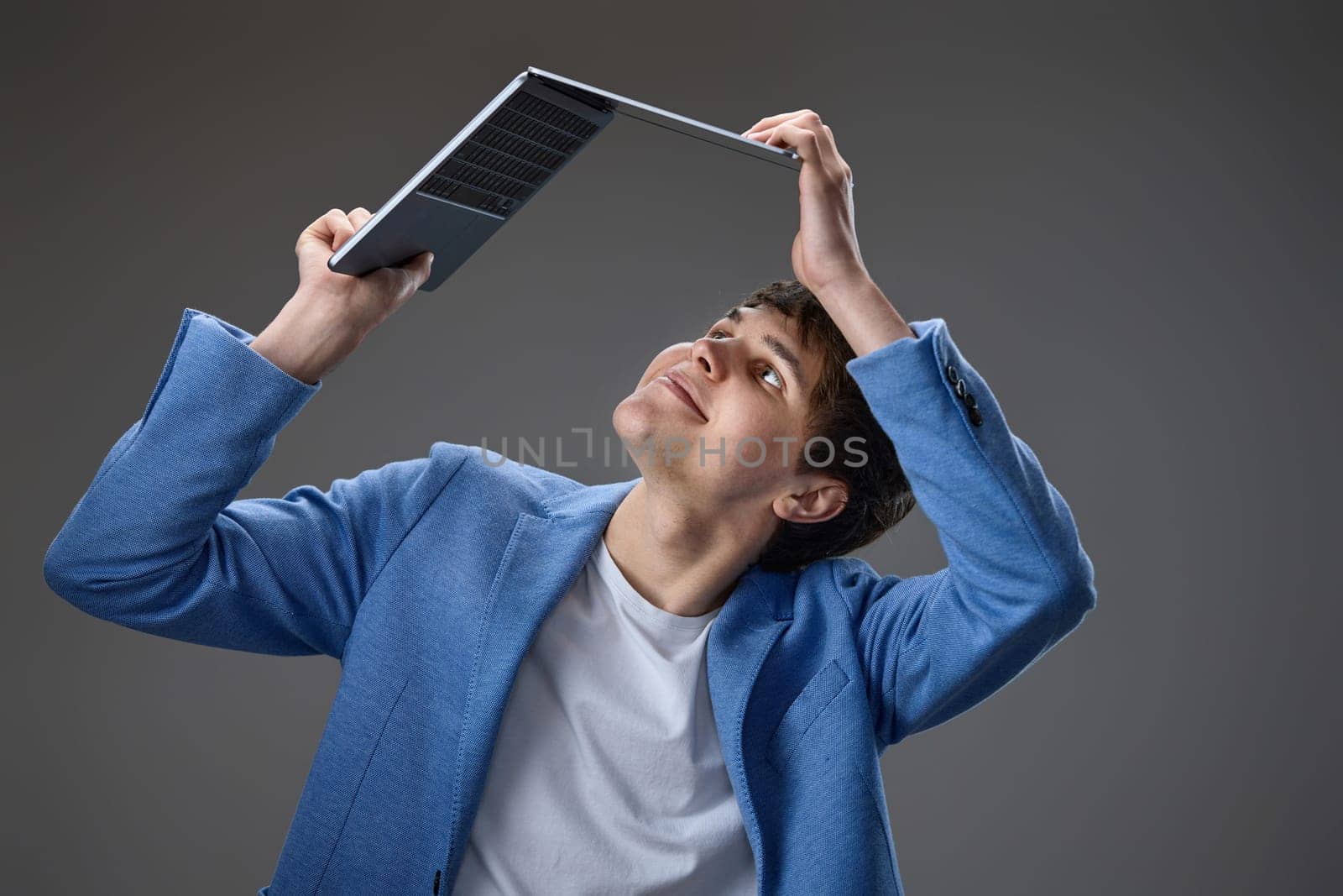 happy caucasian young guy in blue jacket holding laptop above his head on gray background.