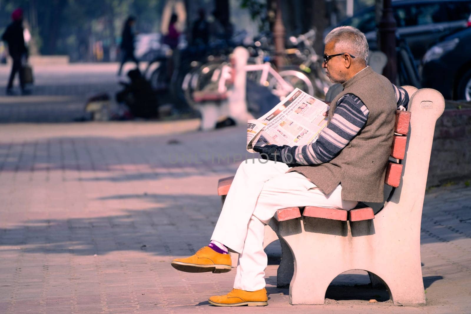 Chandigarh, India - circa 2023 : old man sitting on sunny park bench reading newspaper in sector 17 market in chandigarh, showing this famous landmark shopping area in the city beautiful
