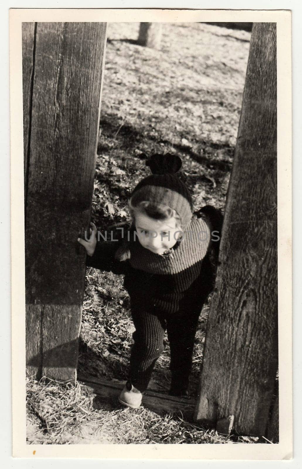 A vintage photo shows small girl goes through the hole in wooden fence. by roman_nerud