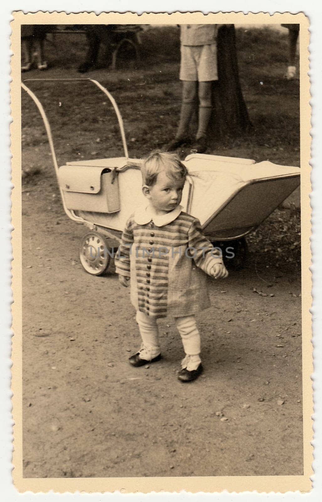 A vintage photo shows small toddler girl and pram (baby carriage). Toddler shows cute face. by roman_nerud