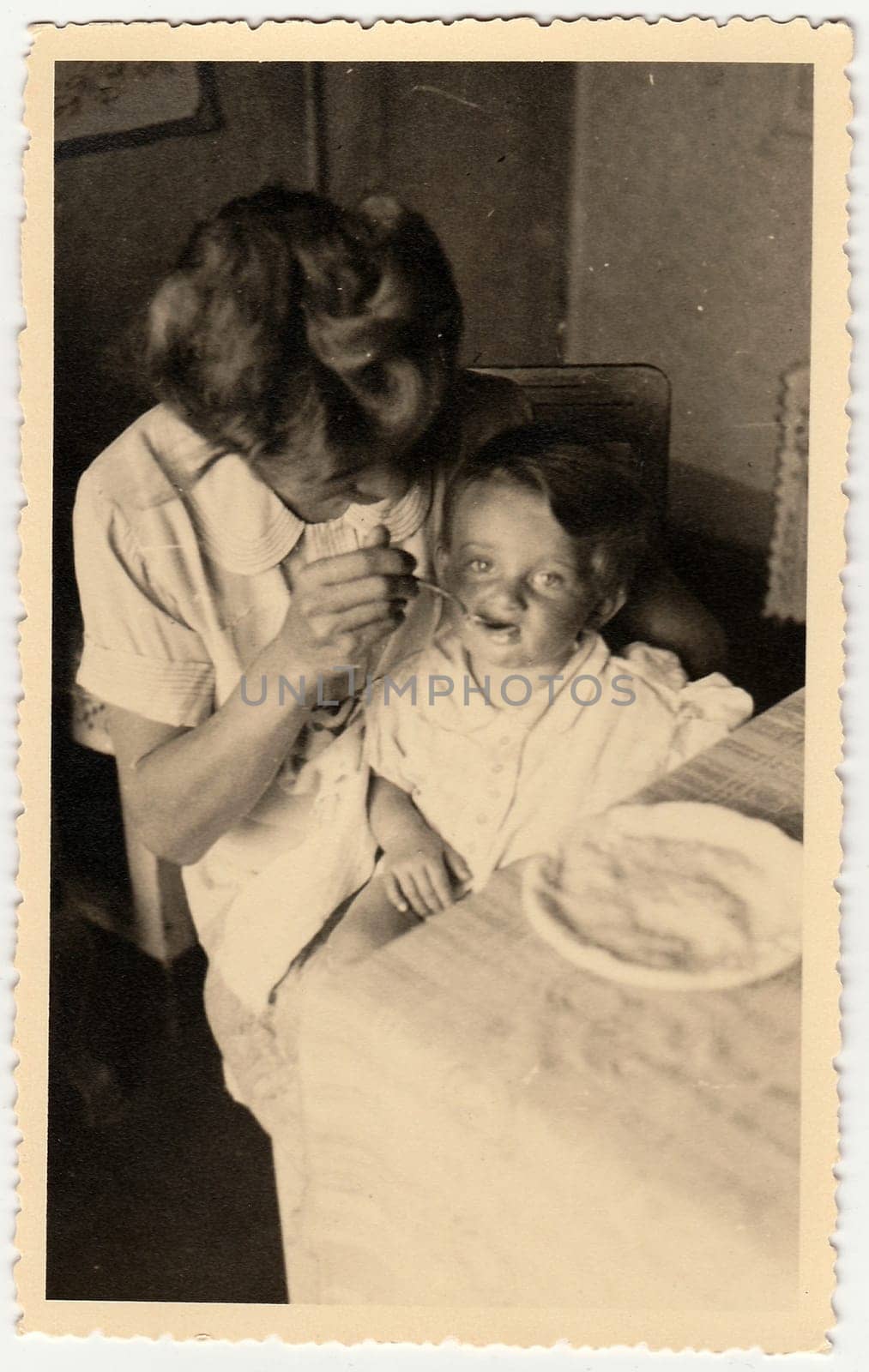 A vintage photo shows small toddler girl and mother feeds her. by roman_nerud