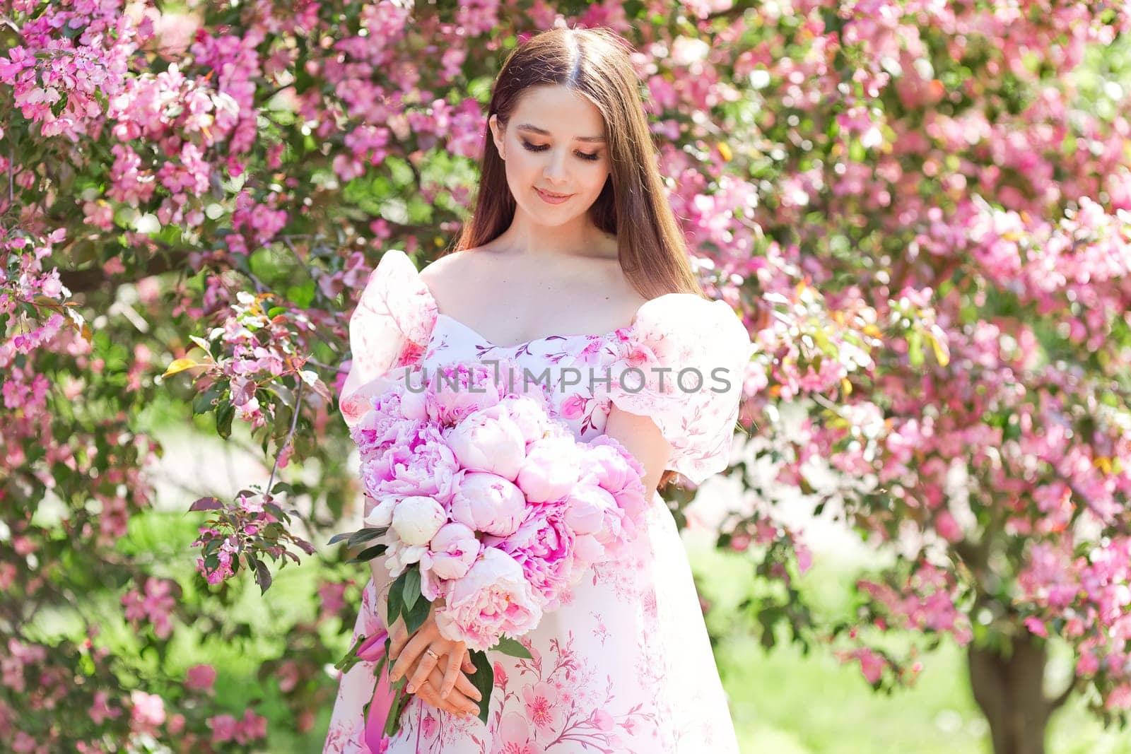 A girl in a light pink dress, with a large bouquet of pink peonies, stands next to her in a pink blooming garden on a sunny day. Copy space