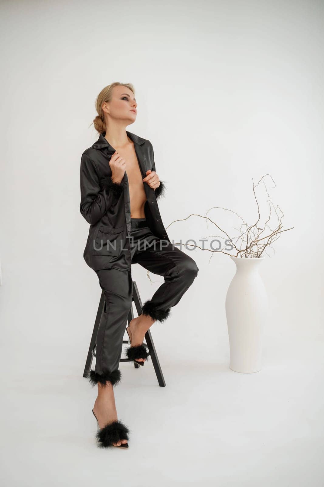 Woman glamorous pajamas. A beautiful blonde in black pajamas sits relaxed on a chair in black pajamas with feathers on a white background. Photo shoot in the studio, advertising pajamas. by Matiunina
