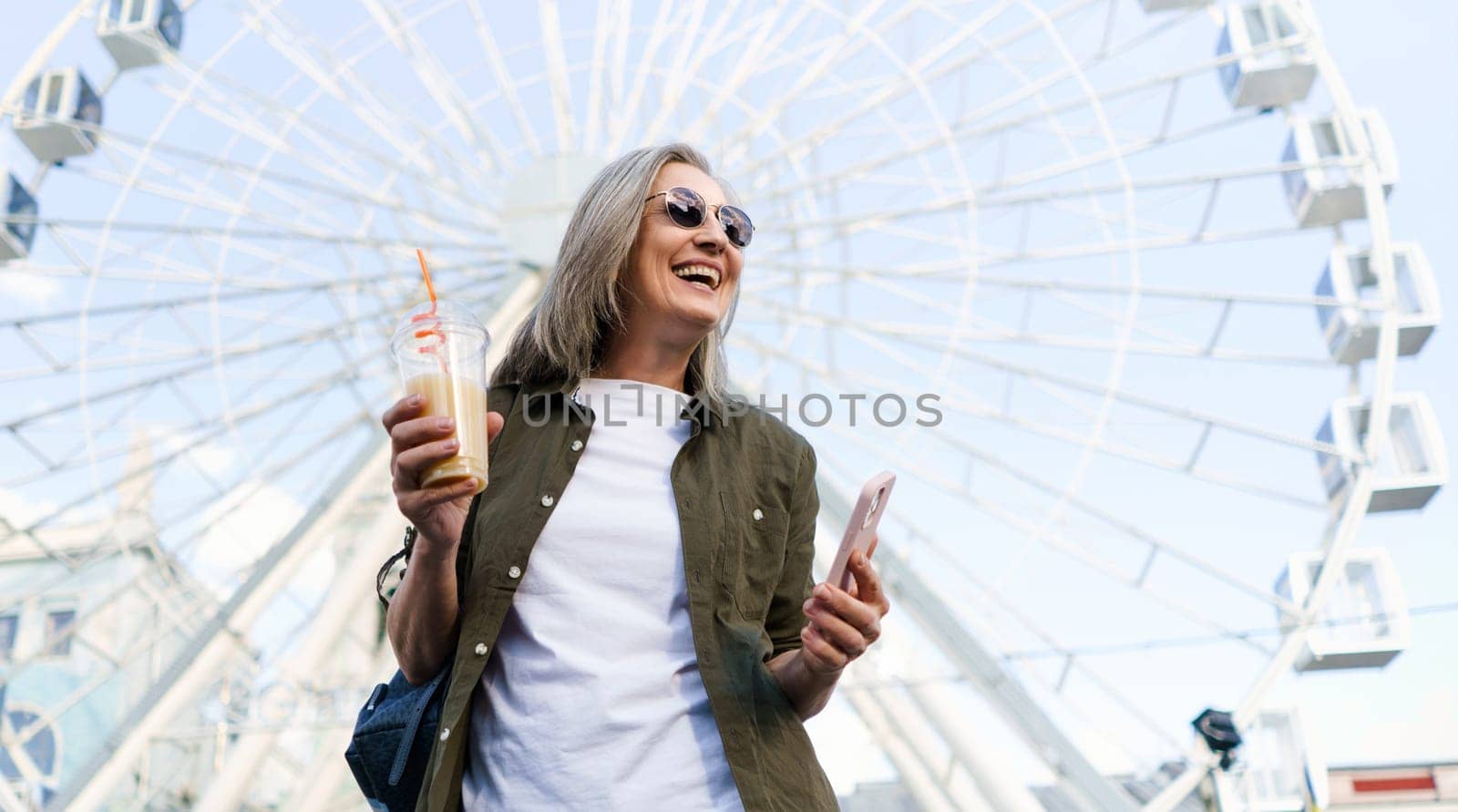 Cheerful senior woman enjoys a sunny day in an old European city, holding her phone and a cup of juice in her hands with a big smile on her face, embodying the concept of elderly happiness. High quality photo