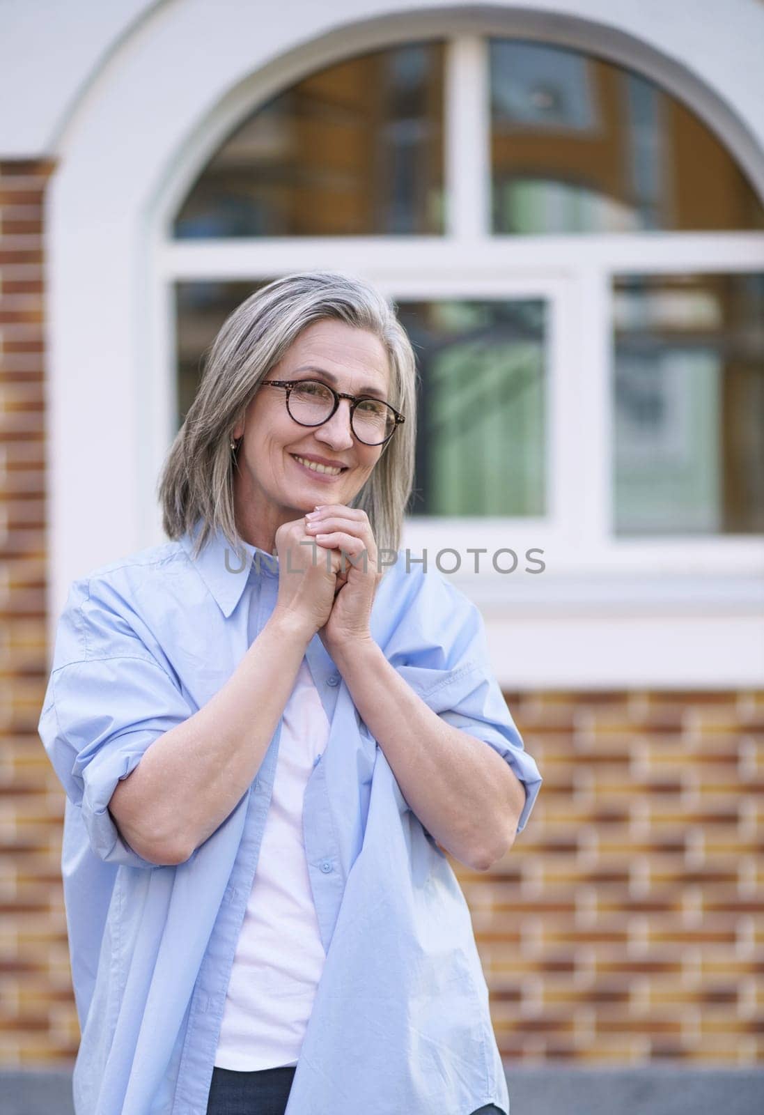 Older woman in a city setting, smiling with her hands clasped together in front of her, conveying a sense of happiness and contentment. High quality photo