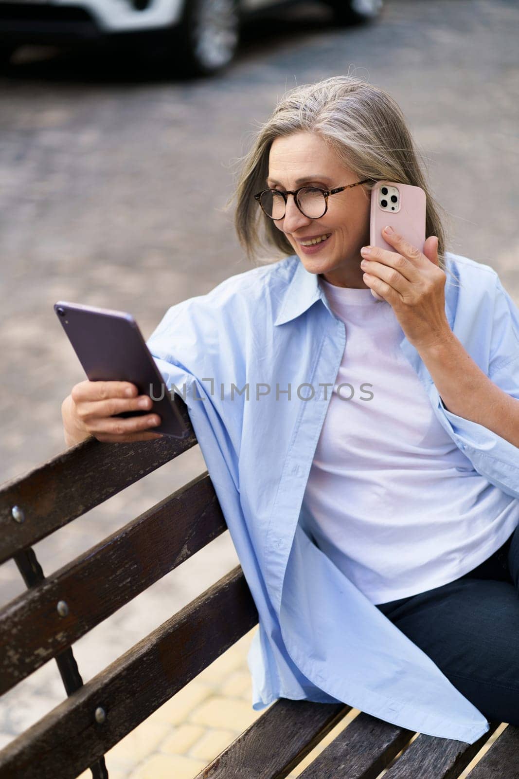 Happy senior woman is multitasking with a tablet PC and phone call on a busy city street. She showcases modern technology and active lifestyle in the elderly population. by LipikStockMedia