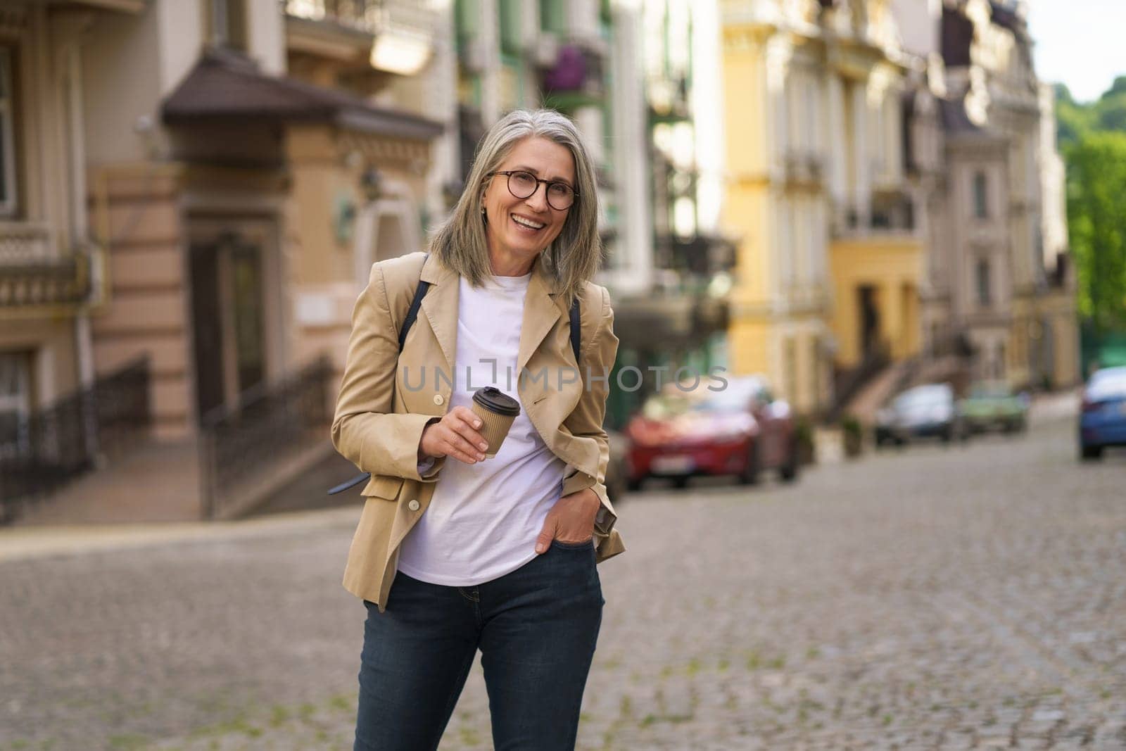 mature woman with a cheerful smile holding a phone while exploring an old European city. The photo captures the concept of elderly happiness and technology use for communication and travel purposes. by LipikStockMedia