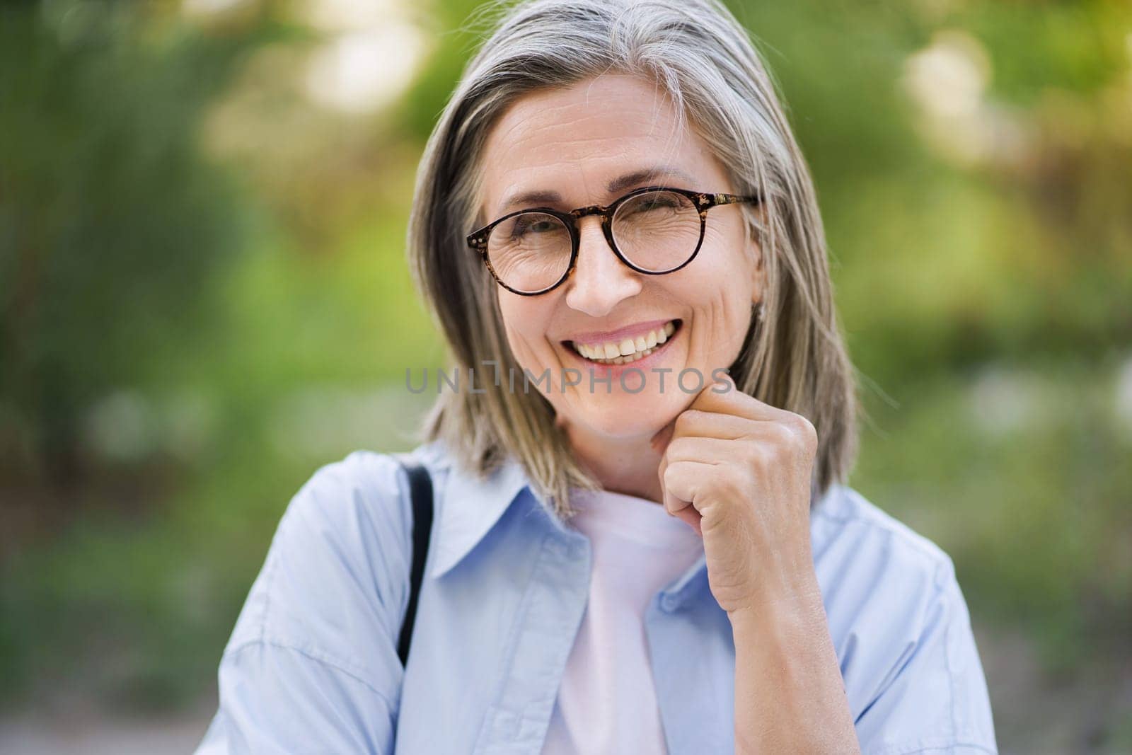 Happy mature woman with silver hair and glasses smiling outdoors, showcasing the beauty of aging and positive attitude towards life in the golden years. by LipikStockMedia