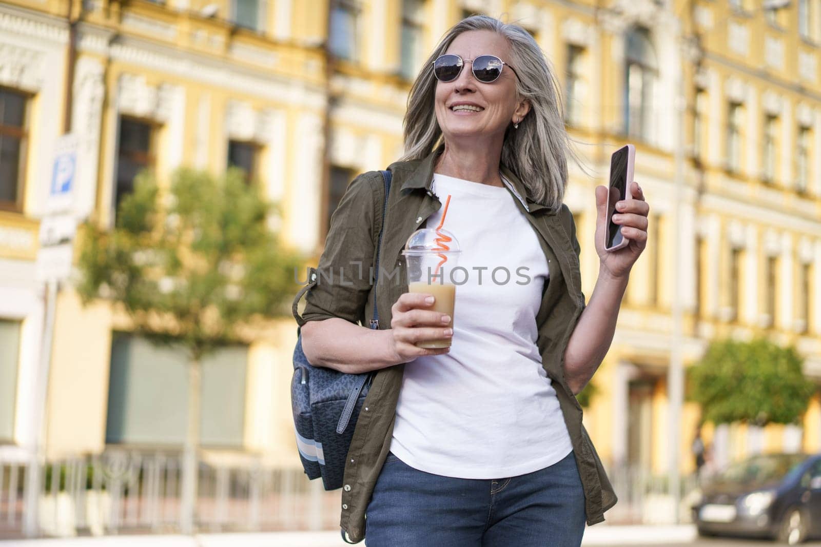 Biohacking Concept. A Mature European Woman with Gray Hair wearing Sunglasses Walks around the City with a Drink and a Smartphone. Healthy Longevity. High quality photo
