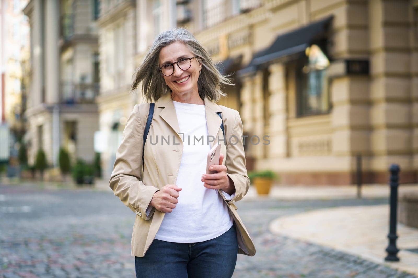 Joyful mature woman holds her phone while standing in old European city. This image captures the concept of happiness in the elderly. by LipikStockMedia