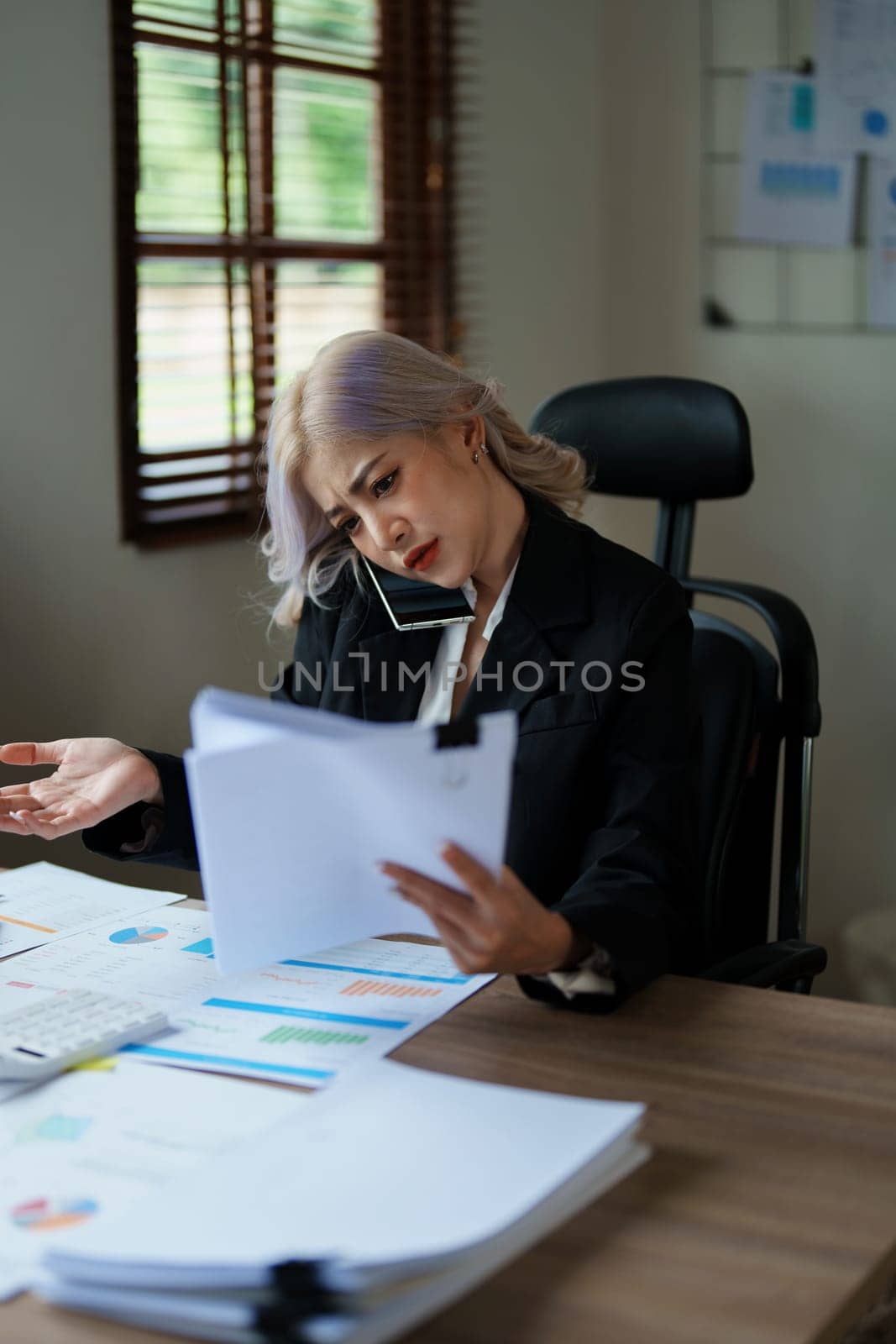 Portrait of a young Asian woman showing a serious face as she uses her phone, financial documents and computer laptop on her desk in the early morning hours.