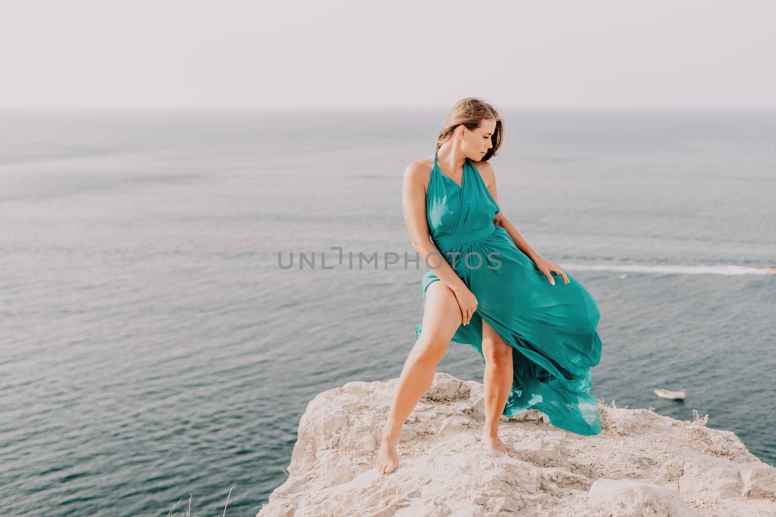 Side view a Young beautiful sensual woman in a mint long dress posing on a volcanic rock high above the sea during sunset. Girl on the nature on overcast sky background. Fashion photo