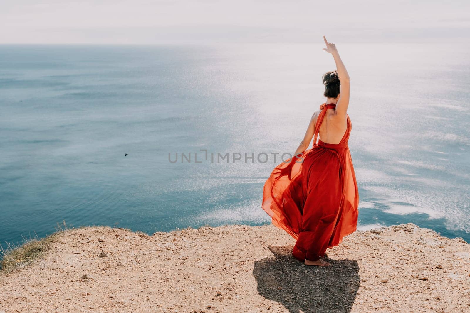 Woman in red dress on sea. Side view a Young beautiful sensual woman in a red long dress posing on a rock high above the sea on sunset. Girl on the nature on blue sky background. Fashion photo. by panophotograph