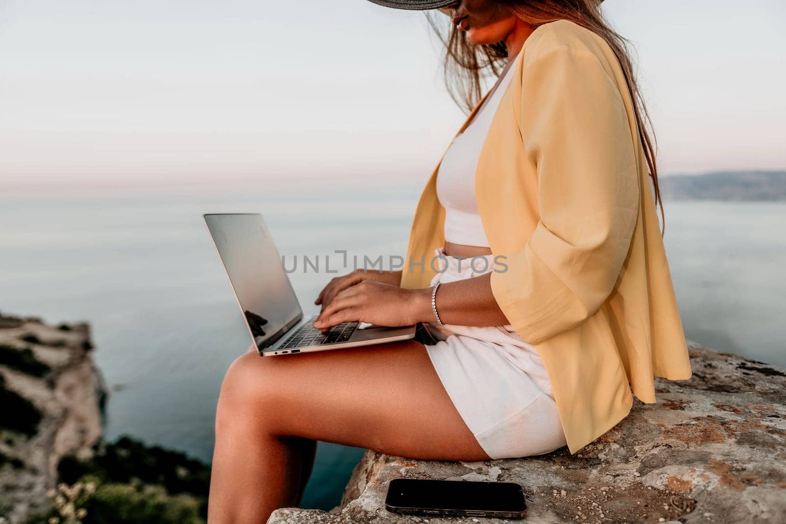 Digital nomad, Business woman working on laptop by the sea. Pretty lady typing on computer by the sea at sunset, makes a business transaction online from a distance. Freelance, remote work on vacation by panophotograph