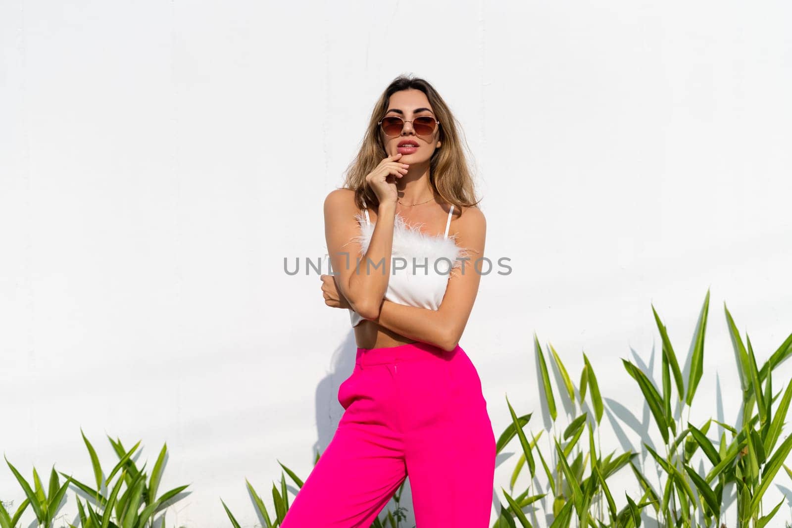Stylish fit tanned beautiful woman in sunglasses, fashion pink pants and white top posing outdoor at white wall tropical leaves background, sunny day, natural light by kroshka_nastya