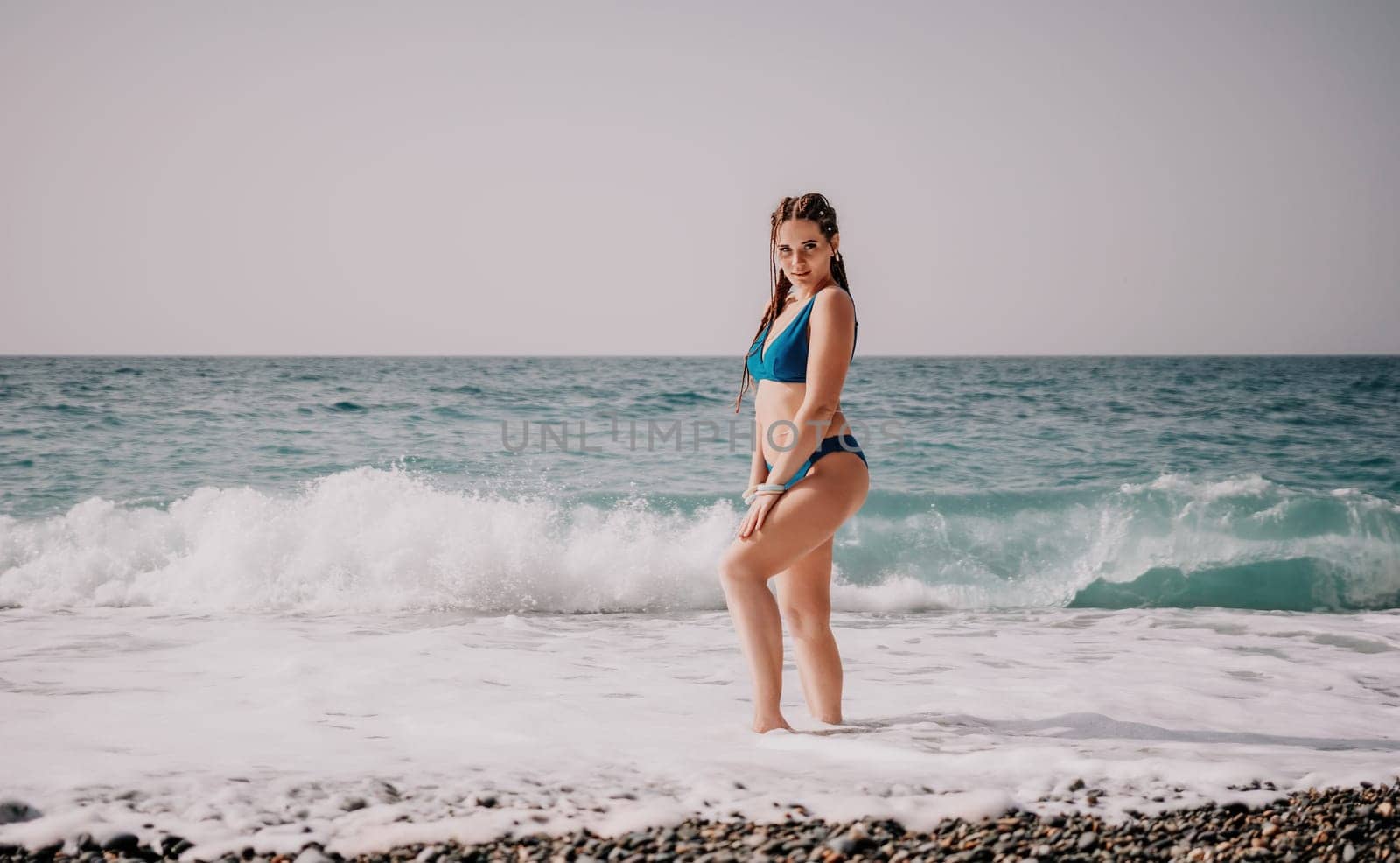 A tourist woman in blue bikini on beach enjoys the turquoise sea during her summer holiday. Rear view. Beach vacation. woman with braids dreadlocks standing with her arms raised enjoying beach ocean. by panophotograph