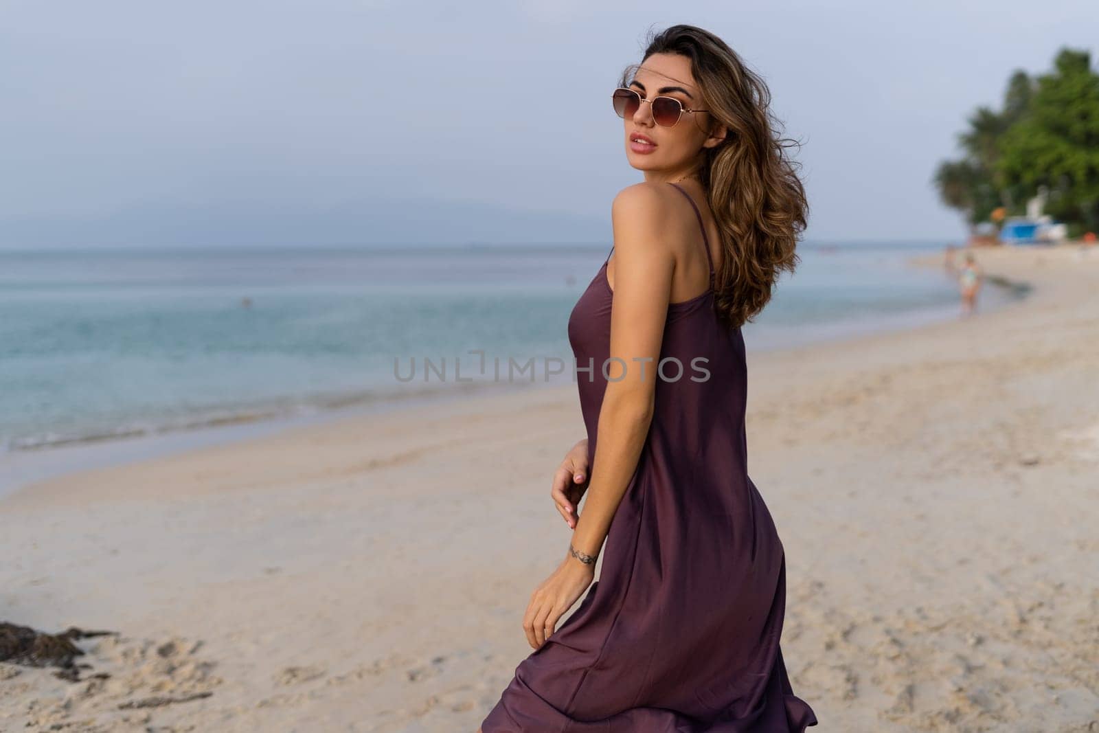 Stylish romantic tender sensual woman in silk dress and sunglasses on the beach at sunset