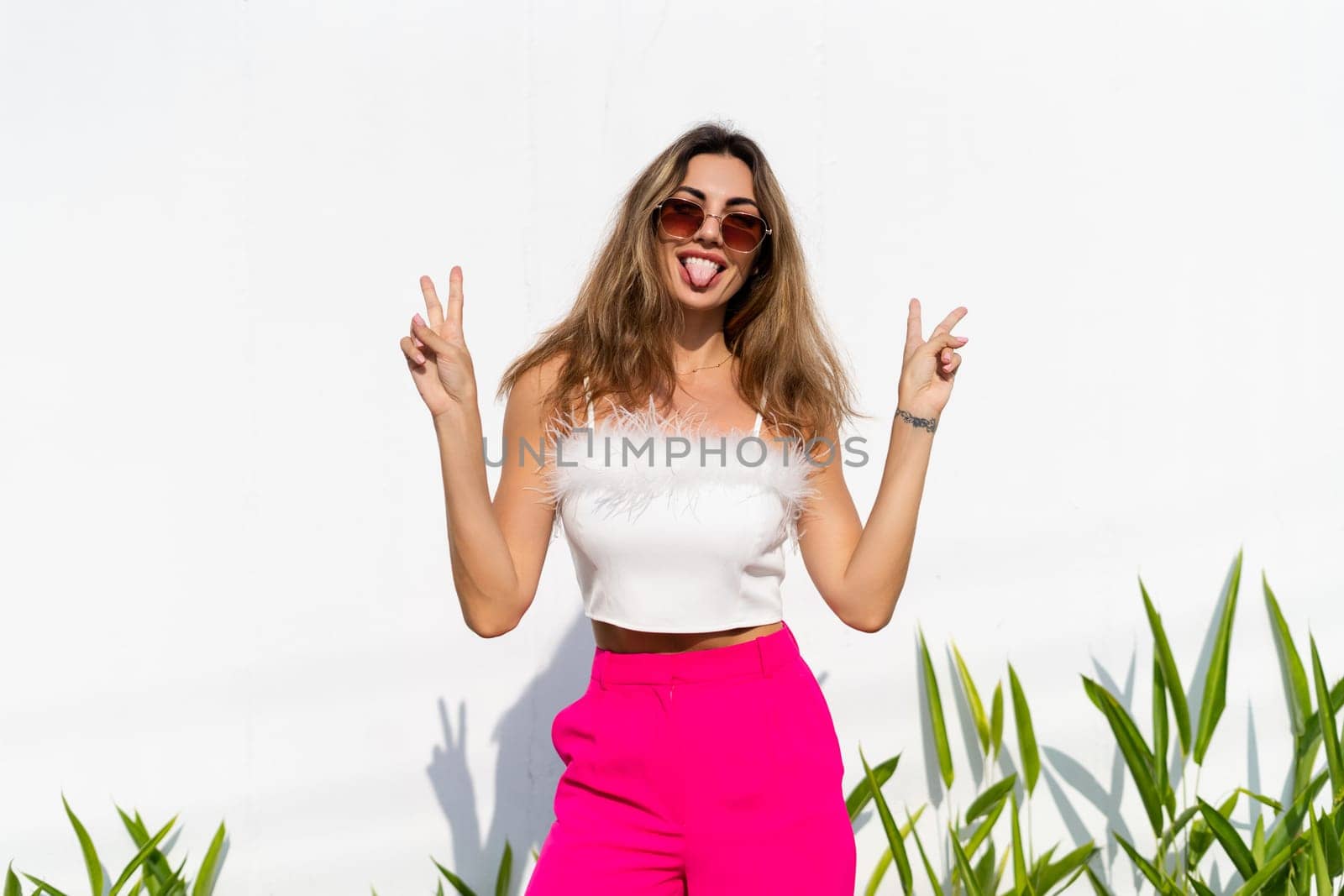 Stylish fit tanned beautiful woman in sunglasses, fashion pink pants and white top posing outdoor at white wall tropical leaves background, sunny day, natural light