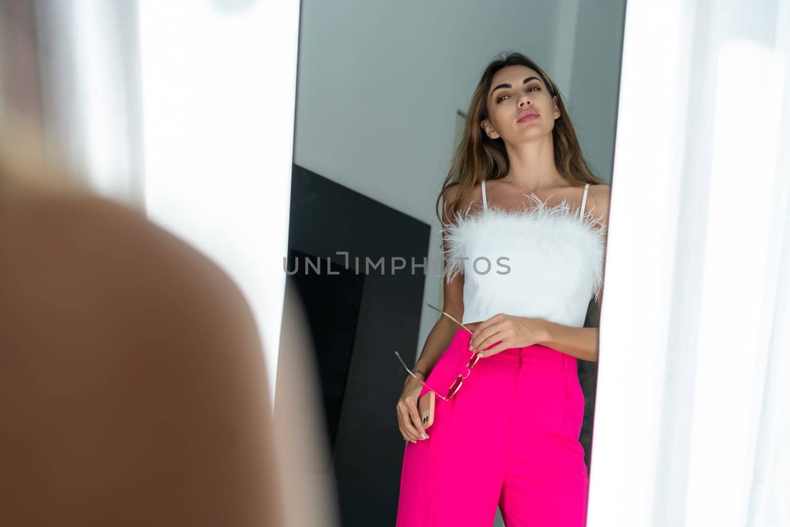Stylish fit tanned beautiful woman in fashion pink pants and top posing in mirror at home bedroom, preparing for party, dressing up
