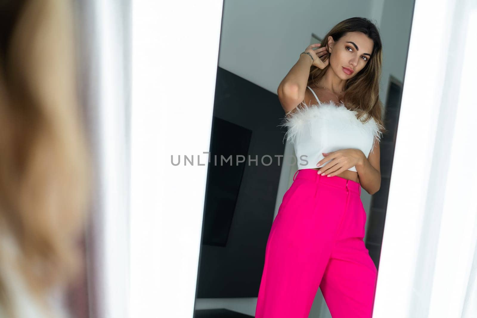 Stylish fit tanned beautiful woman in fashion pink pants and top posing in mirror at home bedroom, preparing for party, dressing up