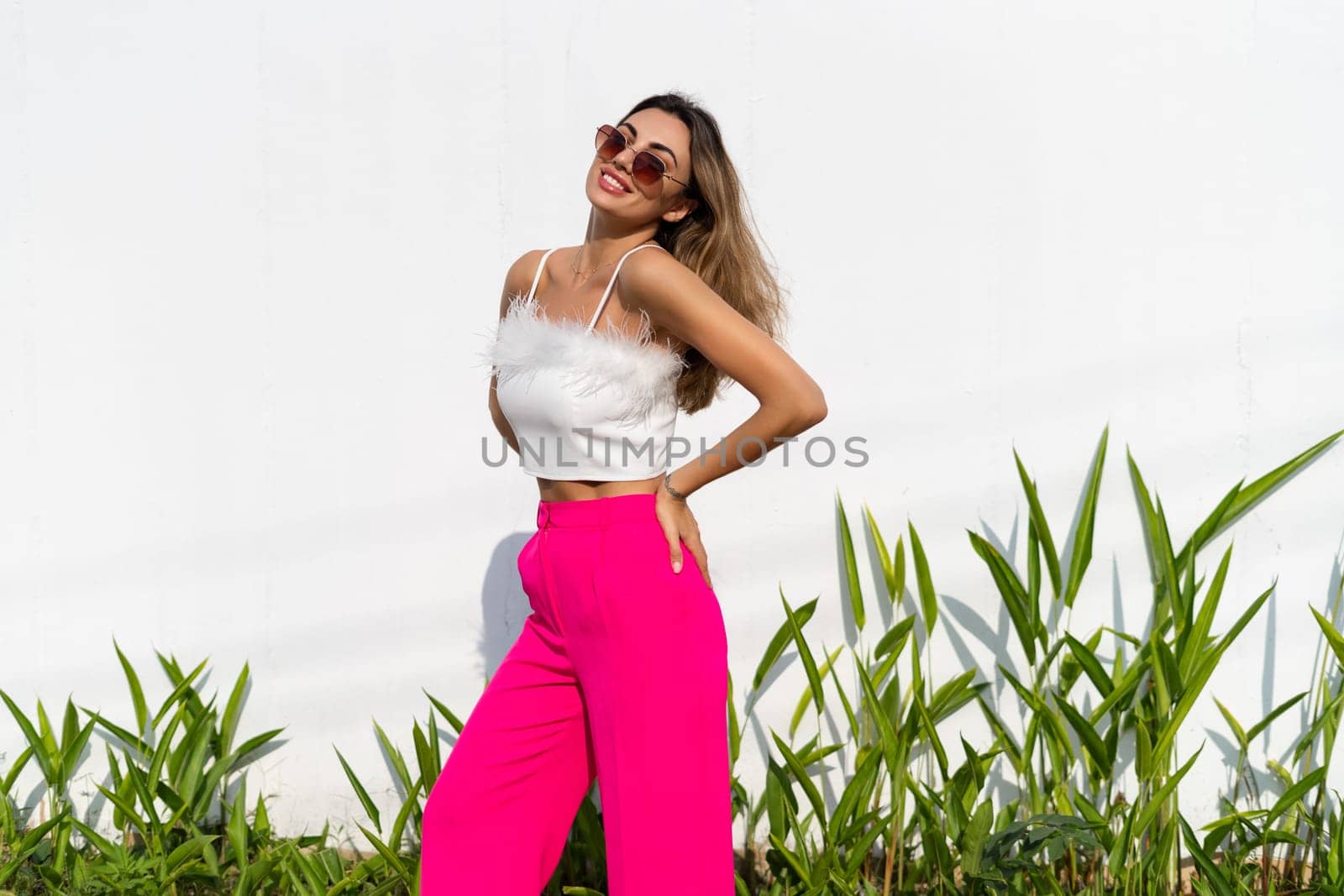 Stylish fit tanned beautiful woman in sunglasses, fashion pink pants and white top posing outdoor at white wall tropical leaves background, sunny day, natural light