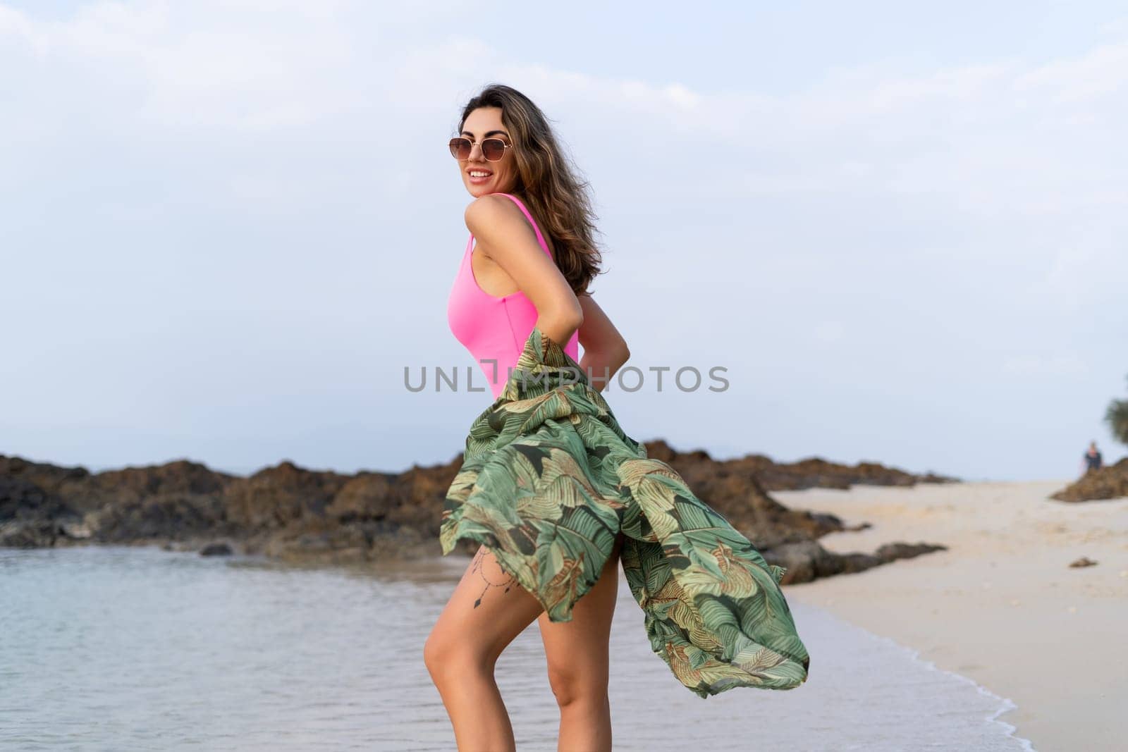 Stylish romantic tender athletic slim sensual woman in pink swimsuit, tropical kimono and sunglasses on the beach at sunset