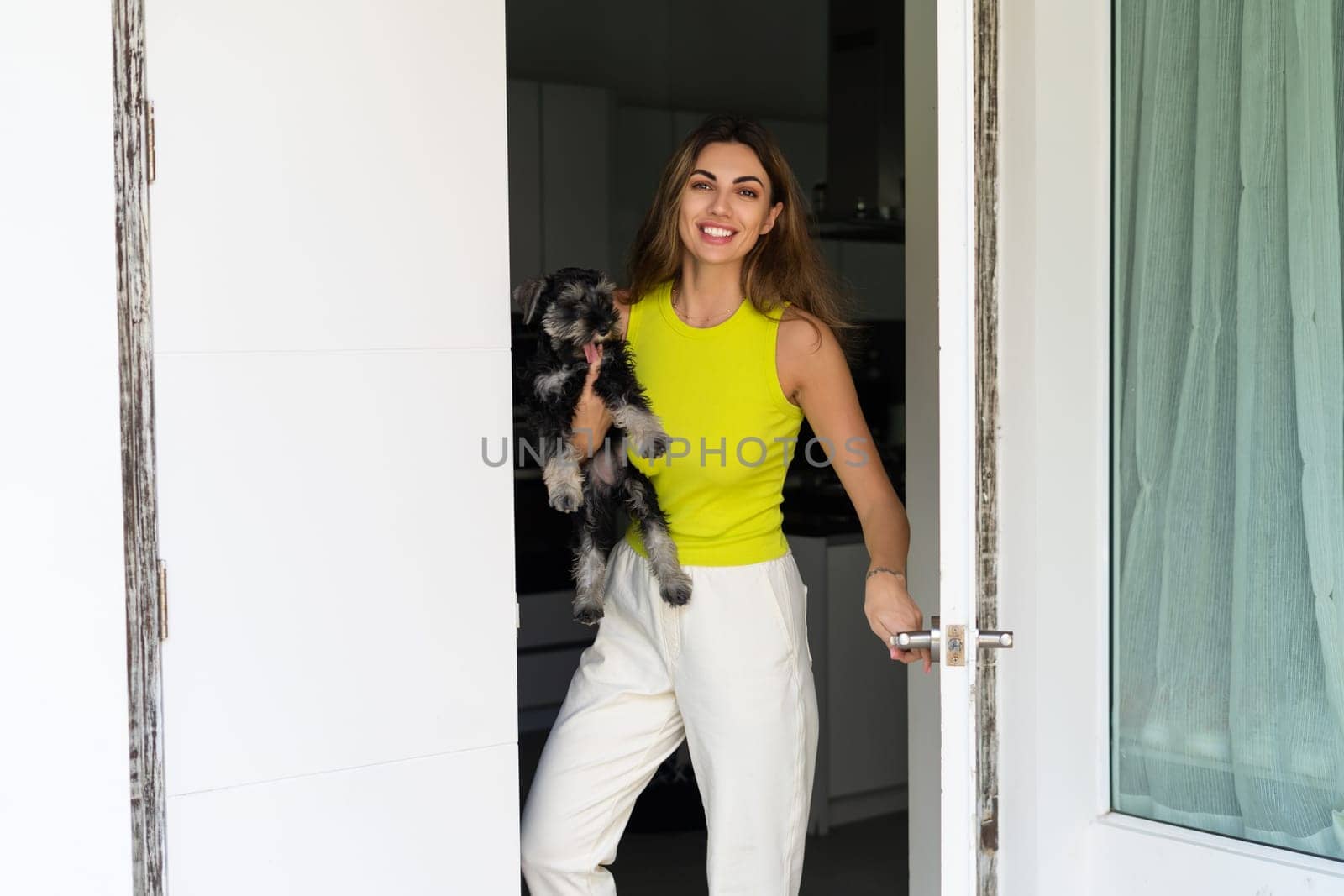 Welcome. Portrait of cheerful woman standing in doorway of modern apartment, greeting visitor and inviting guest to enter her home, holding a puppy