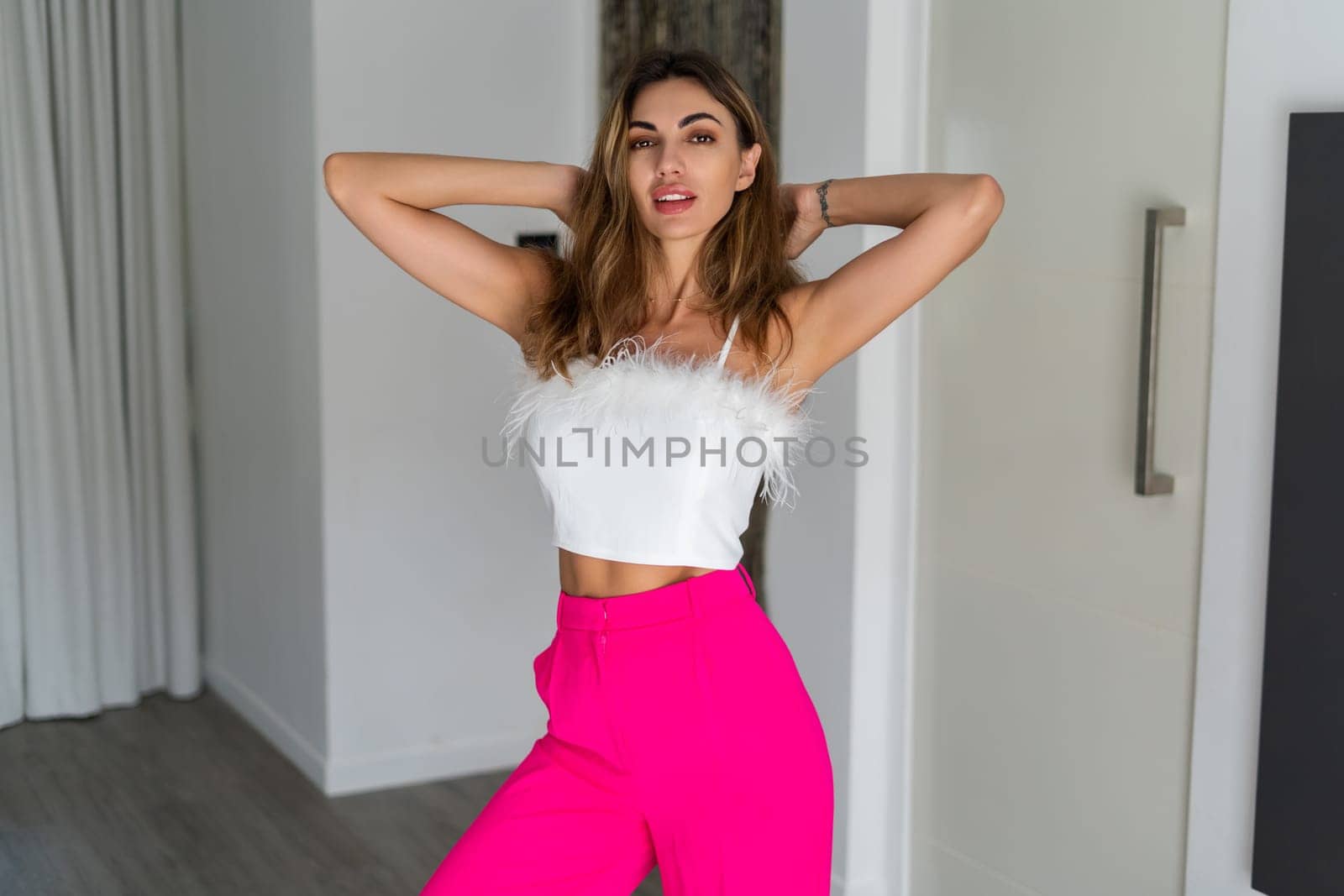 Stylish fit tanned beautiful woman in fashion pink pants and top posing in bedroom at home, preparing for party, dressing up