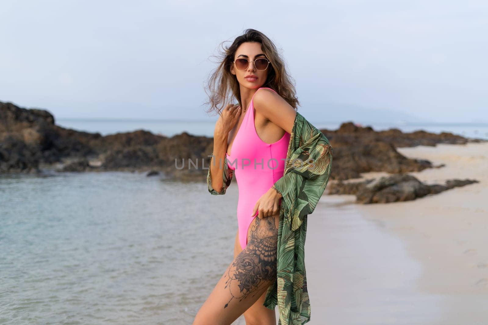Stylish romantic tender athletic slim sensual woman in pink swimsuit, tropical kimono and sunglasses on the beach at sunset