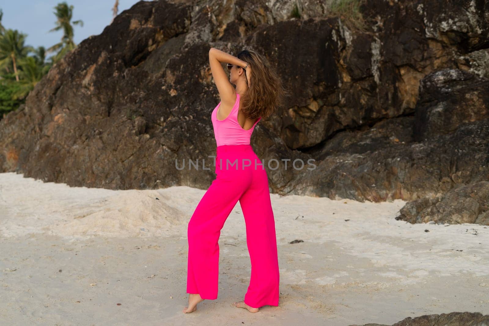 Fashion stylish beautiful sensual young woman, slim, in trendy bright pink pants and bodysuit on a tropical beach at sunset posing in sunglasses