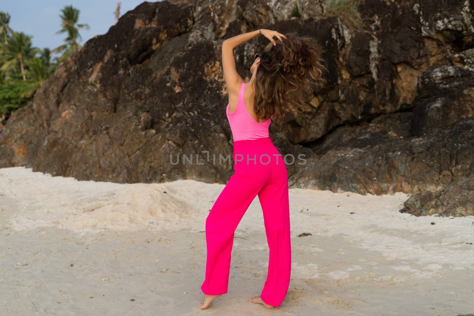 Fashion stylish beautiful sensual young woman, slim, in trendy bright pink pants and bodysuit on a tropical beach at sunset posing in sunglasses