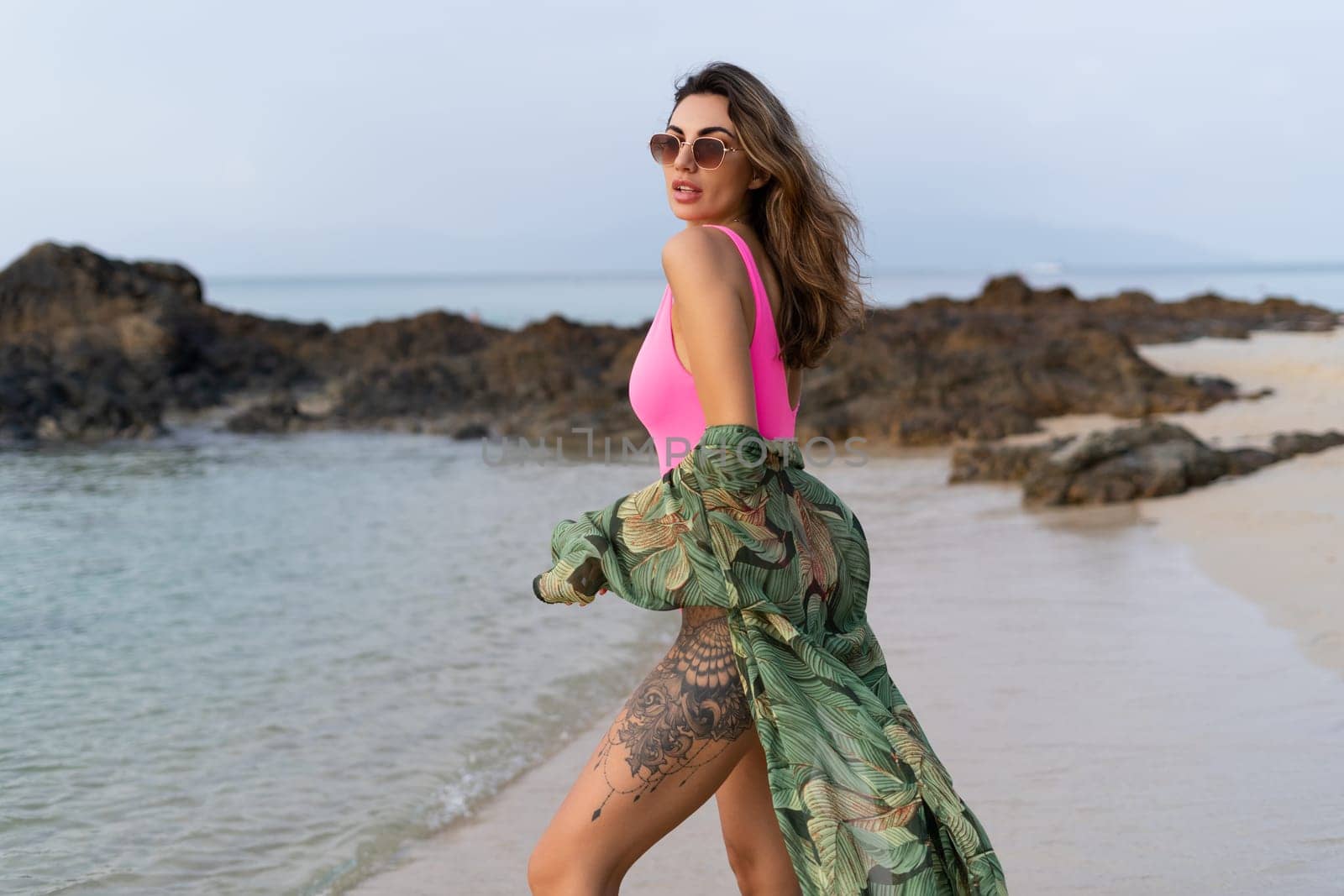 Stylish romantic tender athletic slim sensual woman in pink swimsuit, tropical kimono and sunglasses on the beach at sunset