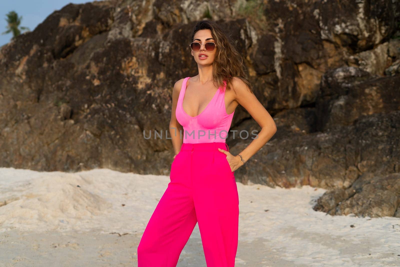 Fashion stylish beautiful sensual young woman, slim, in trendy bright pink pants and bodysuit on a tropical beach at sunset posing in sunglasses