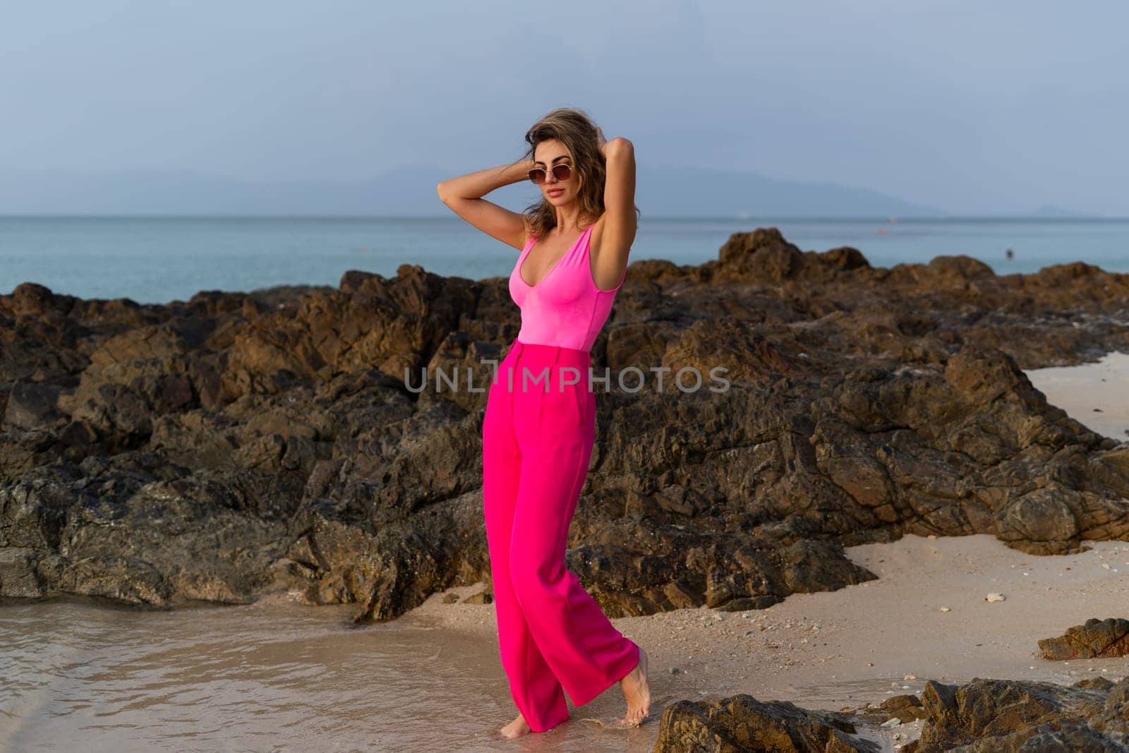 Fashion stylish beautiful sensual young woman, slim, in trendy bright pink pants and bodysuit on a tropical beach at sunset posing in sunglasses