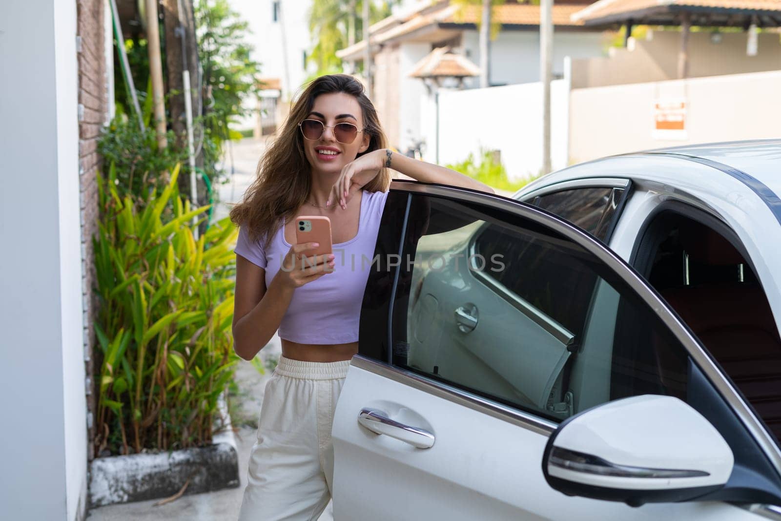 Successful smiling fit tanned attractive woman in casual wear is using her smart phone while standing near luxury modern car outdoors.