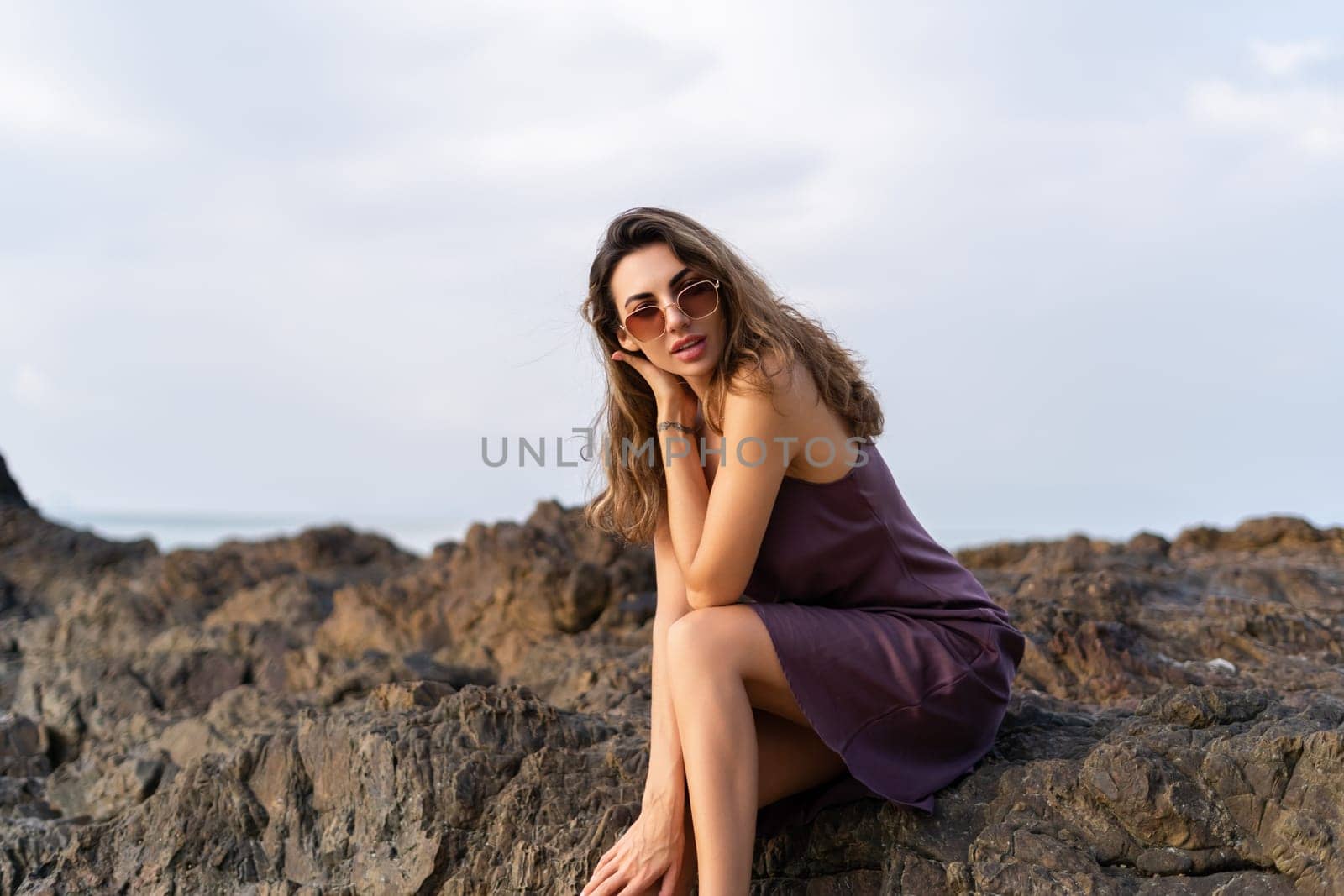 Stylish romantic tender sensual woman in silk dress and sunglasses on the beach at sunset