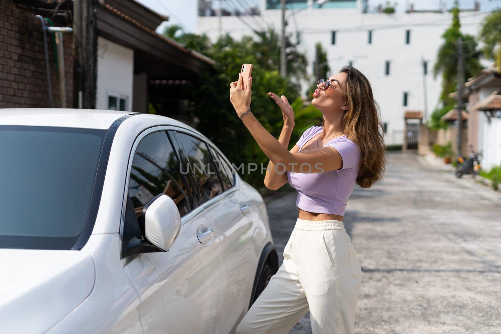 Successful smiling fit tanned attractive woman in casual wear is using her smart phone while standing near luxury modern car outdoors. by kroshka_nastya